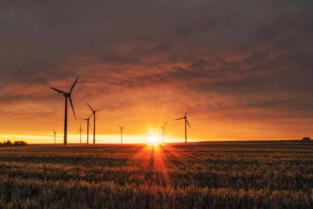 Wind turbines in a field at sunrise, with vibrant orange sky and sunbeams illuminating the landscape.